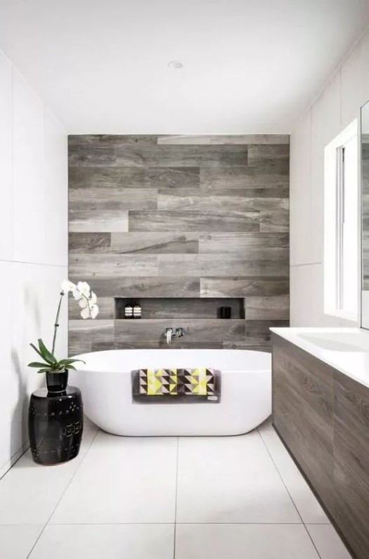 a modern refined bathroom clad with neutral and wood look tiles, with a niche, an oval tub, a wooden vanity and potted blooms