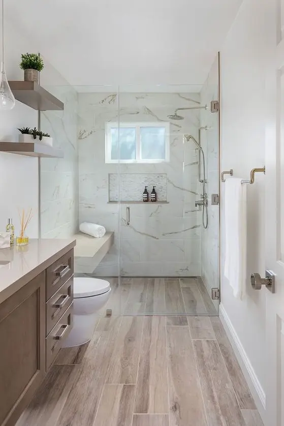 a pretty contemporary bathroom clad with white stone tiles and wood look ones on the floor, a wooden vanity, open shelves and cool brass fixtures